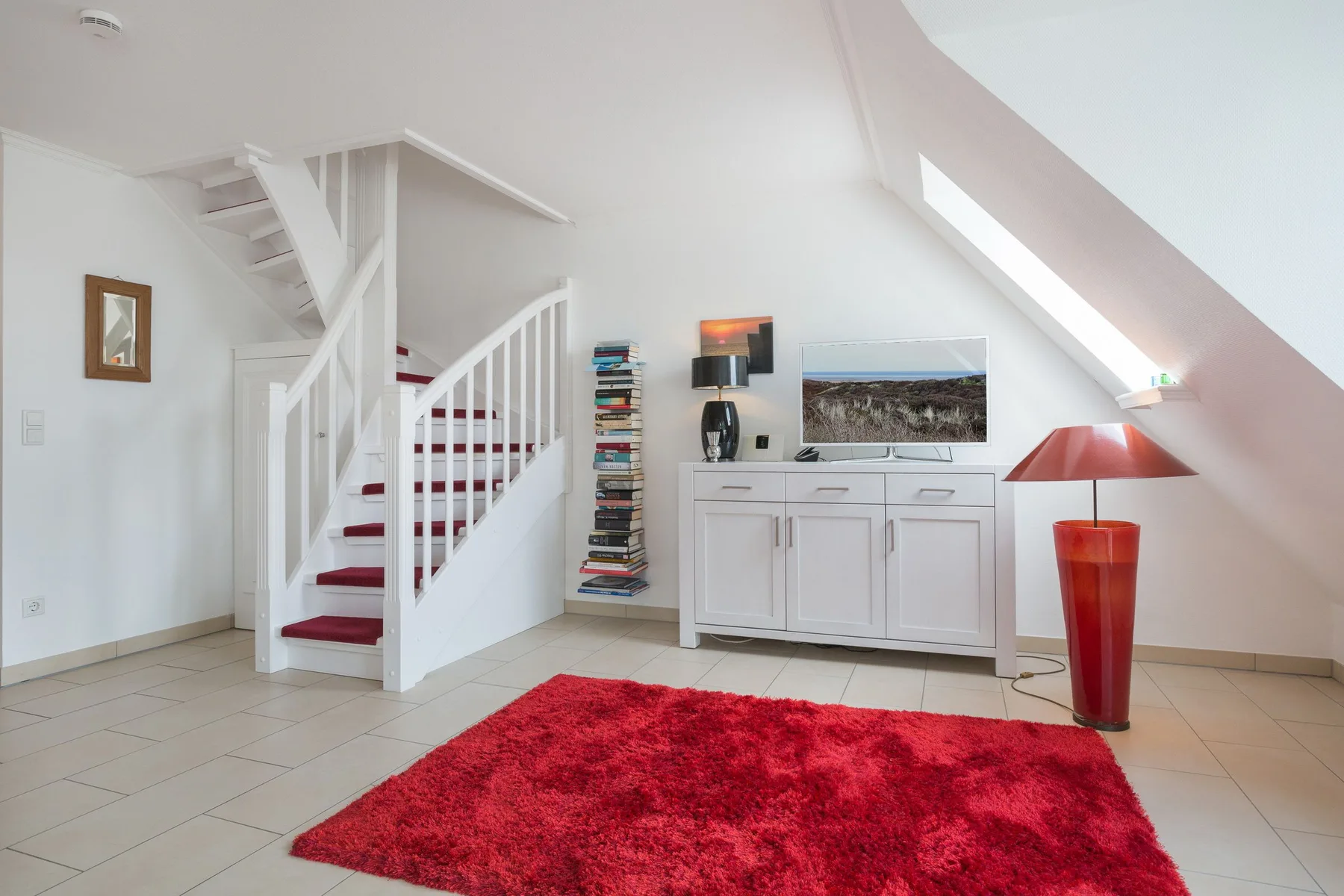 Un luminoso espacio abuhardillado con escalera, mueble blanco y alfombra roja.