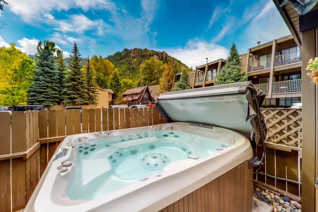 An outdoor hot tub sits beside a wooden deck, with wooded hills and buildings beyond.