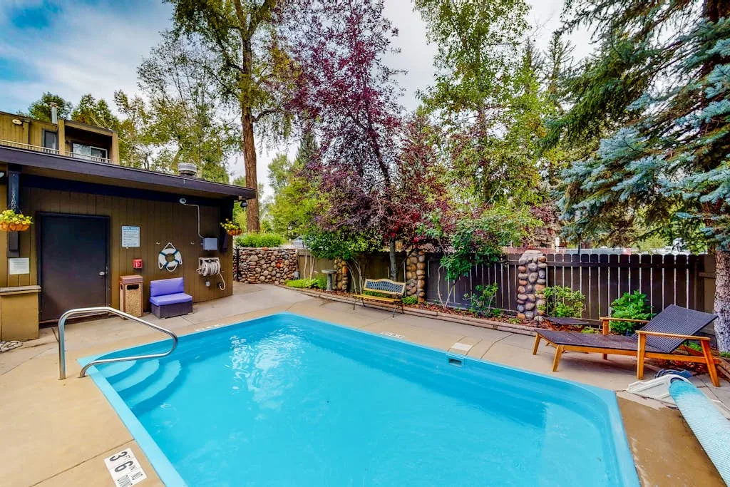 A backyard swimming pool is surrounded by trees, fencing, and outdoor seating.