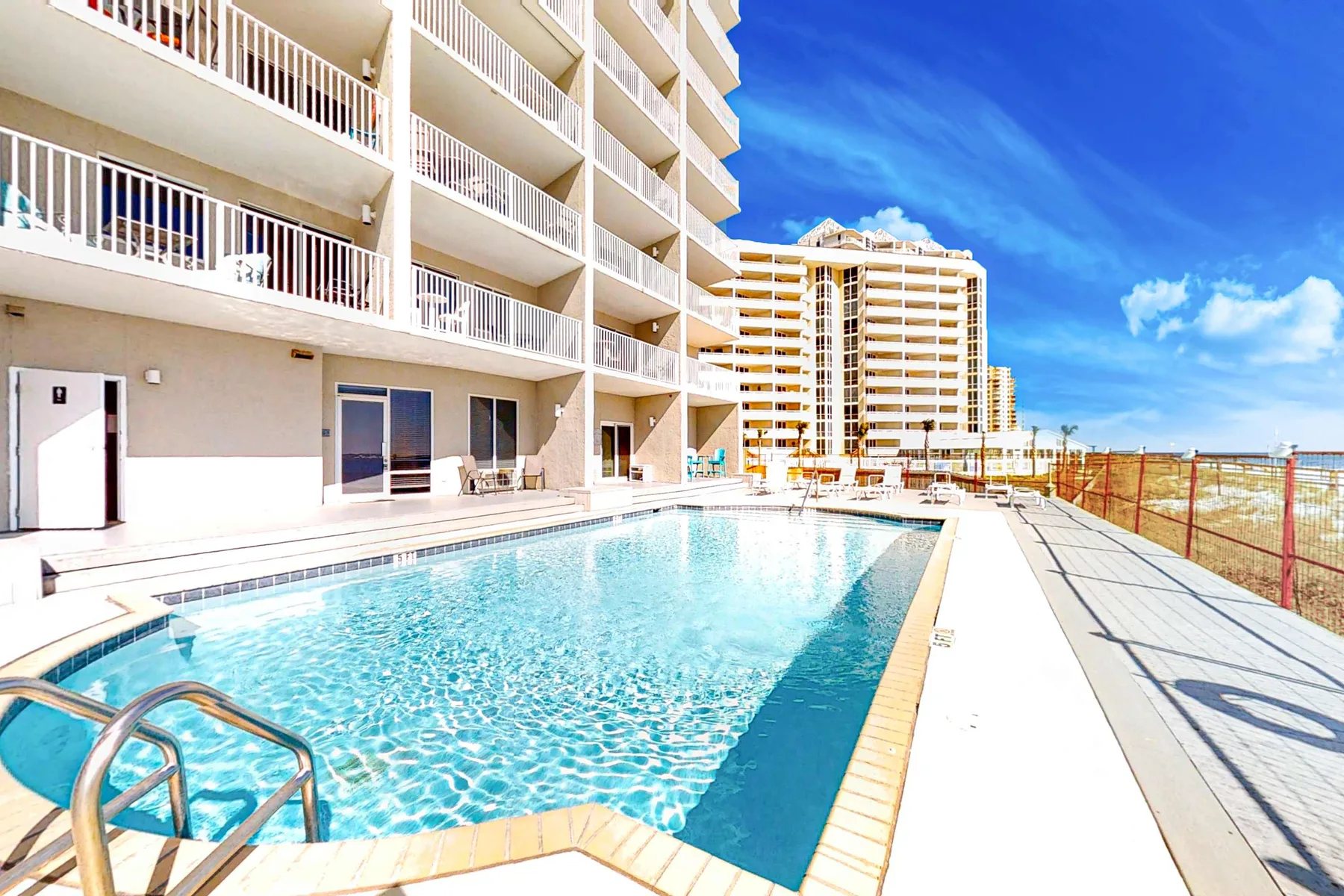 Una piscina al aire libre junto a un edificio de varios pisos bajo un cielo azul despejado.