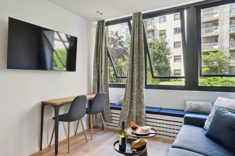 A bright living area with a sofa, wall-mounted television, and large windows.