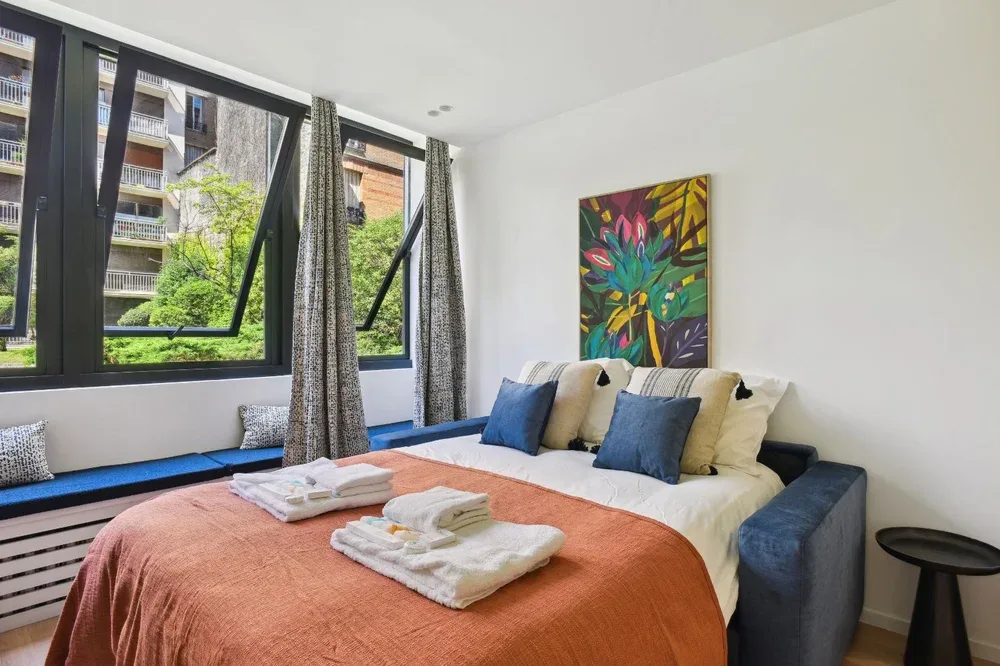 A bright bedroom with a bed beneath colorful wall art and large windows.
