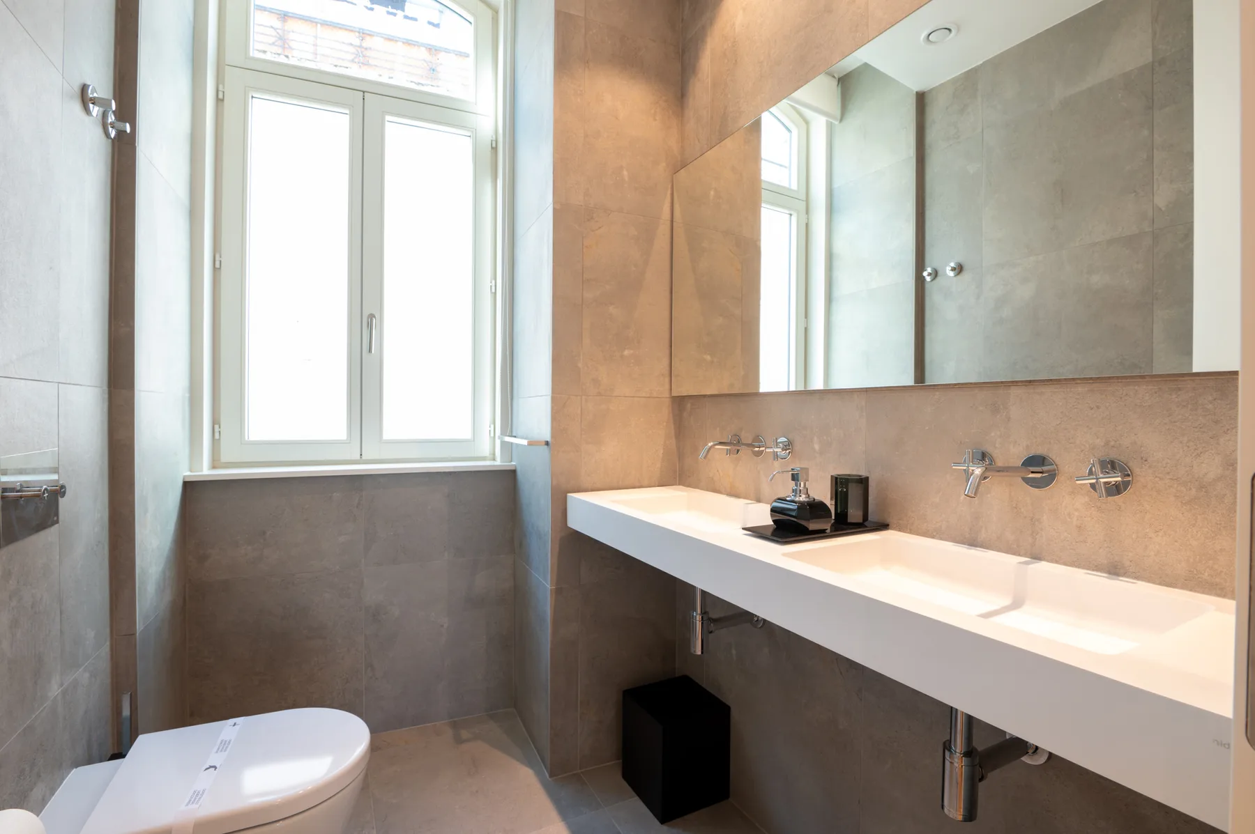 A bathroom with a long white sink counter, large mirror, and window.
