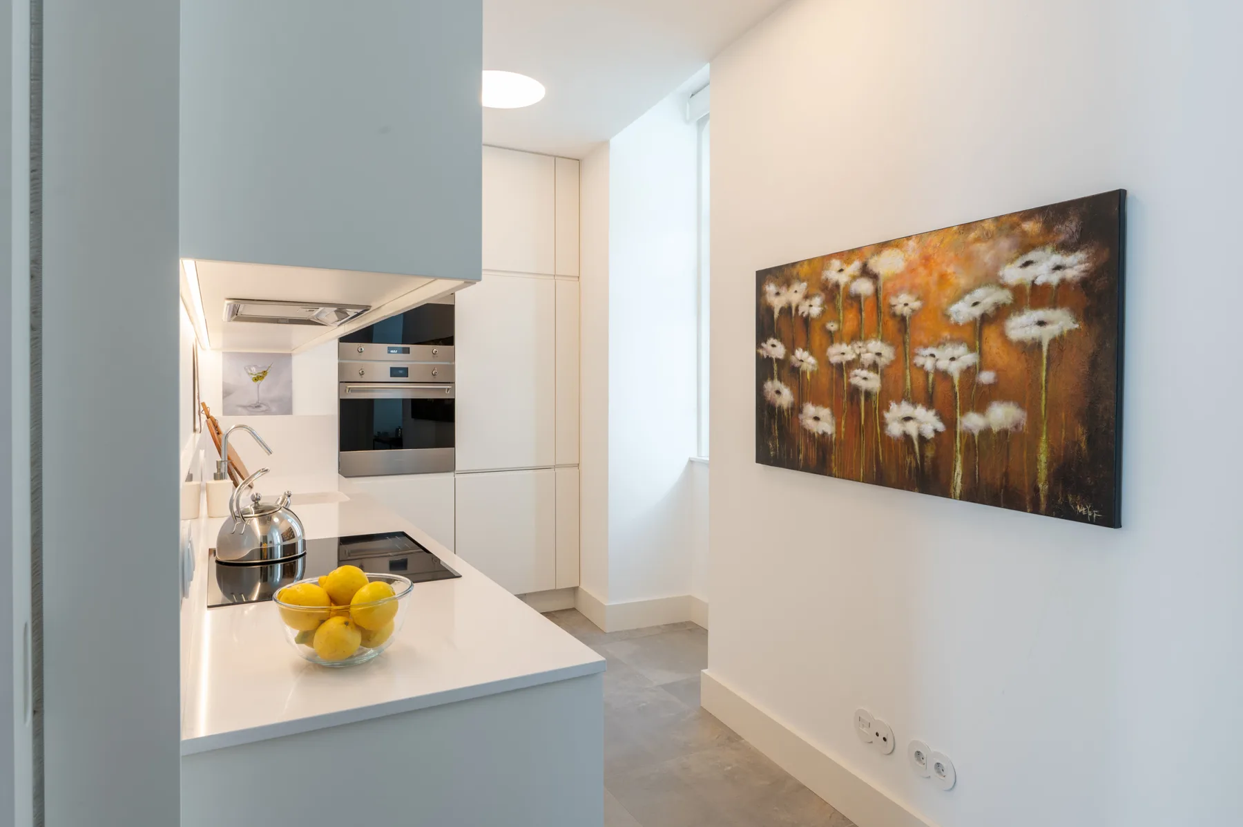 Una cocina luminosa con una encimera blanca y un cuadro floral en la pared.