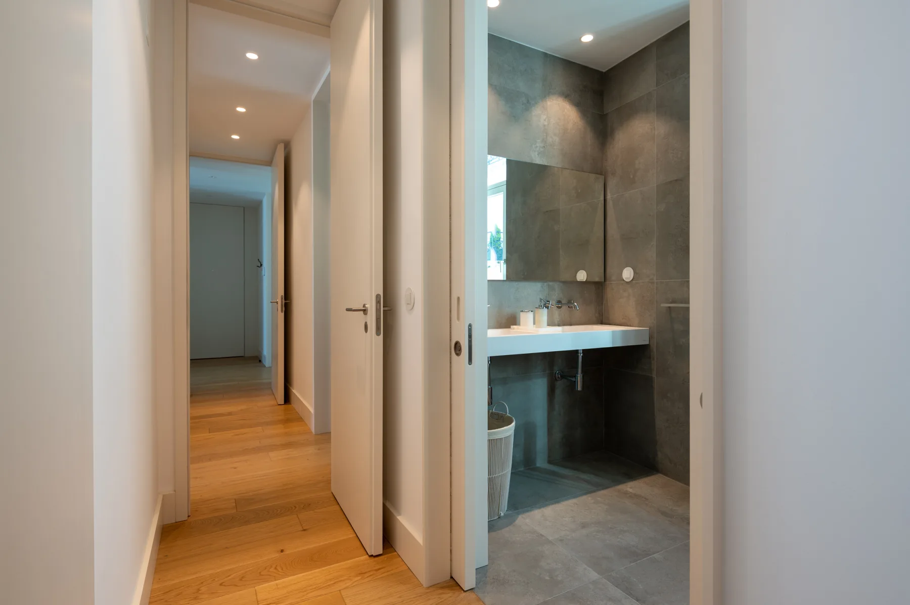 A hallway with wood flooring leads to a bathroom with a sink and shower.