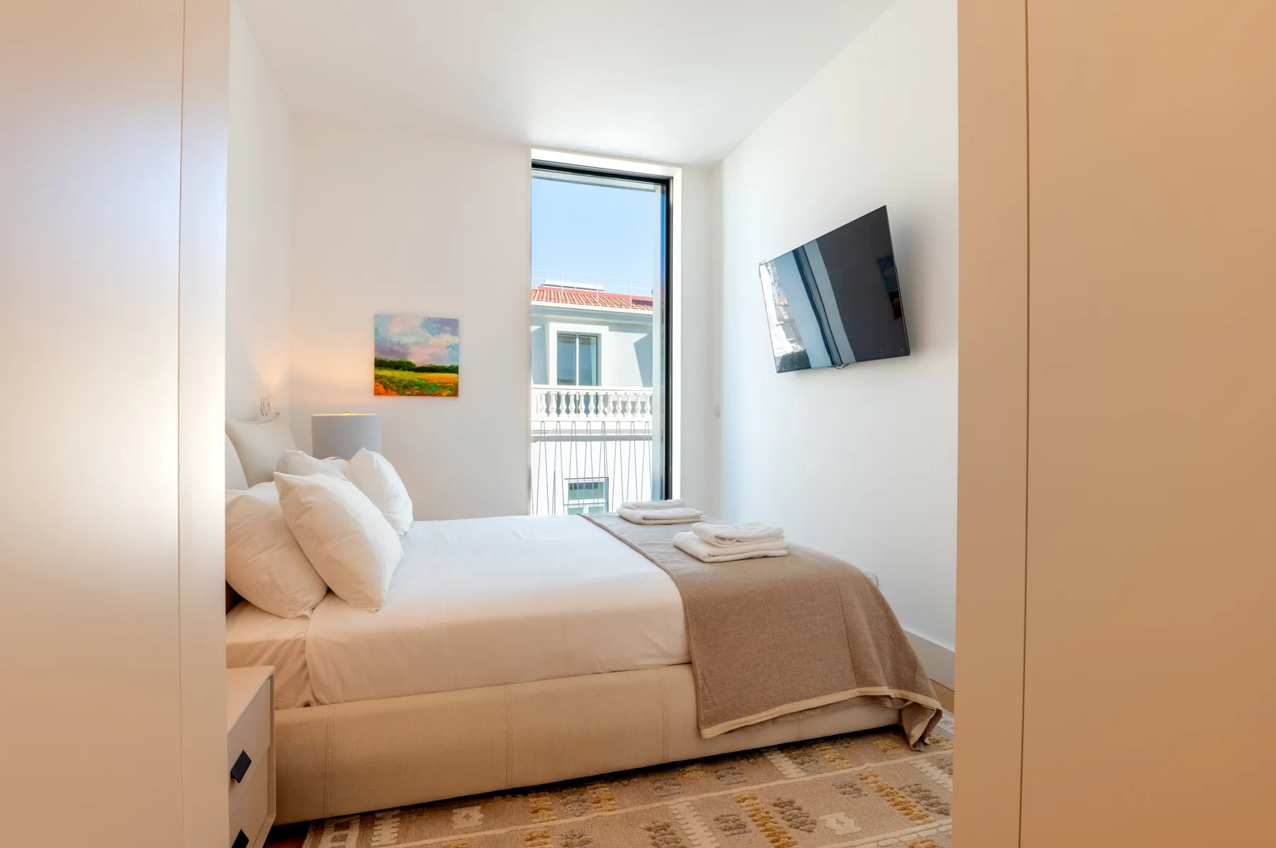 A bright bedroom with a bed and a window overlooking nearby buildings.