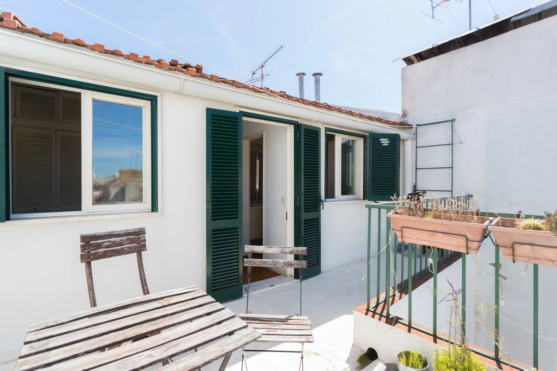 Una terraza soleada con asientos de madera y contraventanas verdes junto a la puerta.