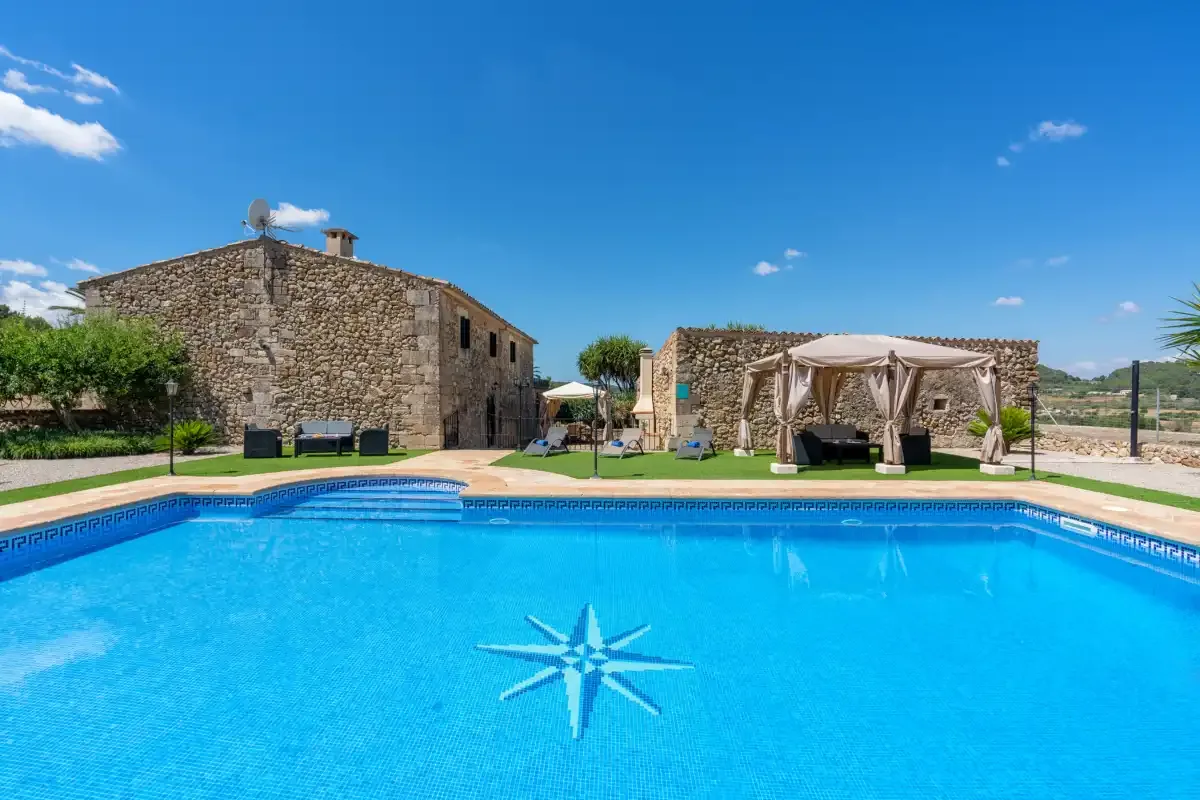 A swimming pool with a star design sits beside stone buildings and a shaded seating area.