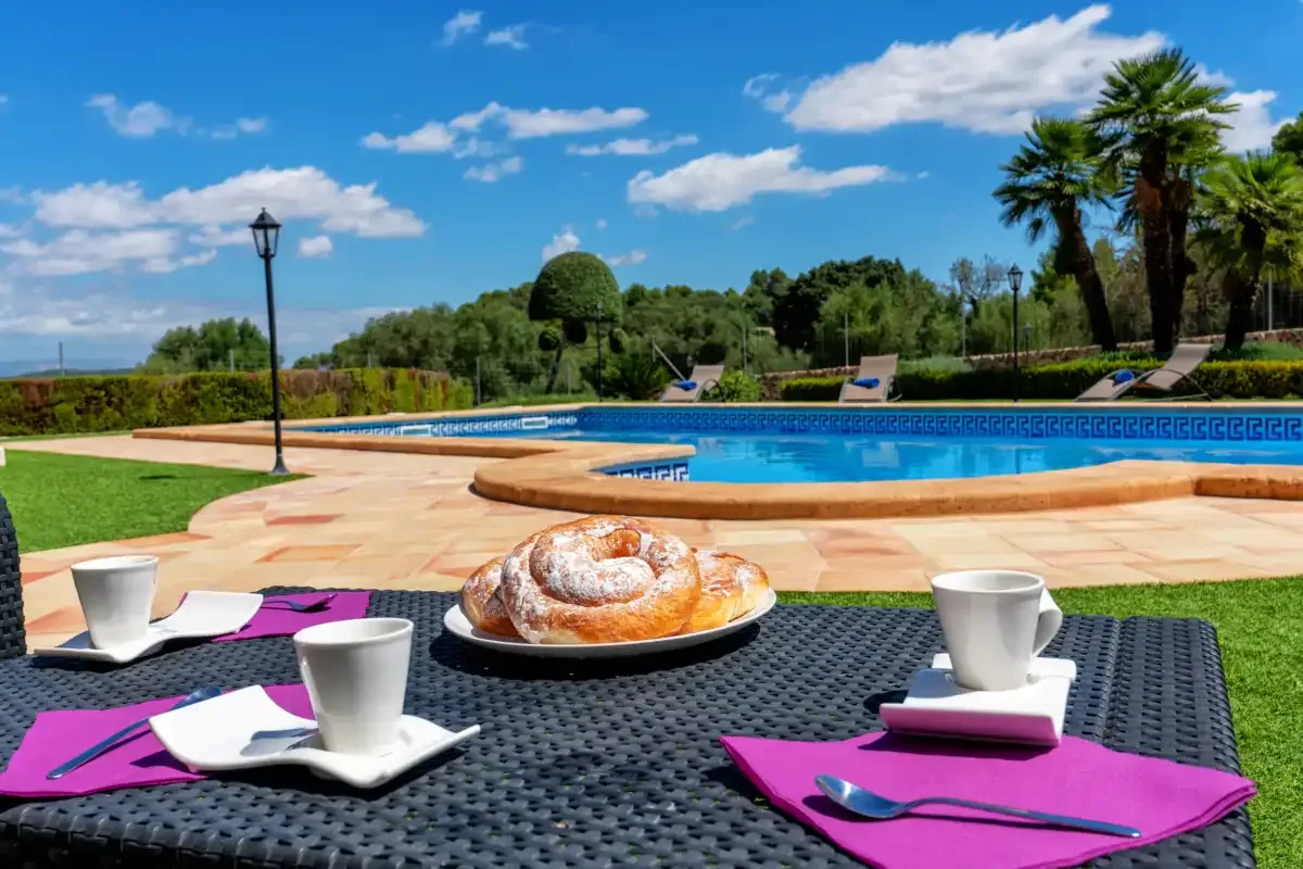 A poolside patio table is set with pastries and cups beside a swimming pool.