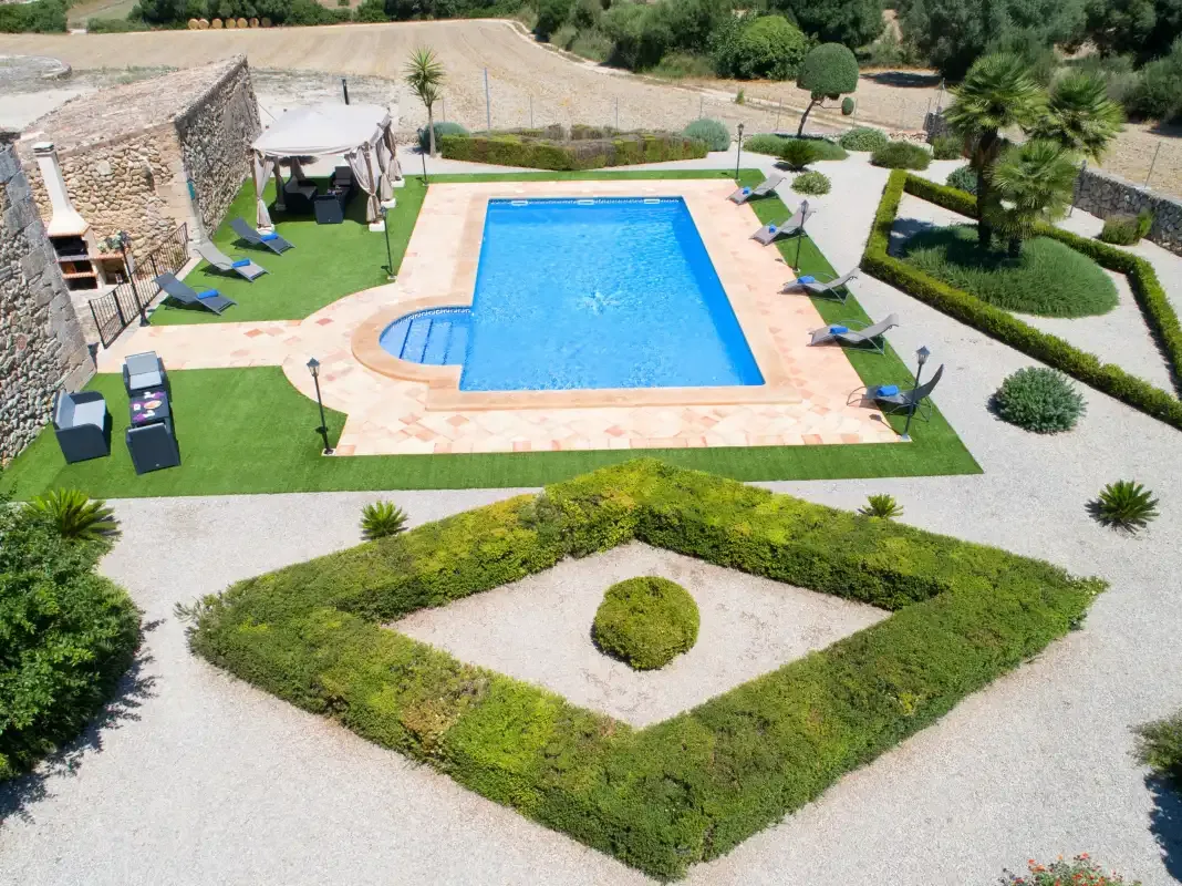 An outdoor swimming pool is surrounded by paved areas, greenery, and lounge seating.