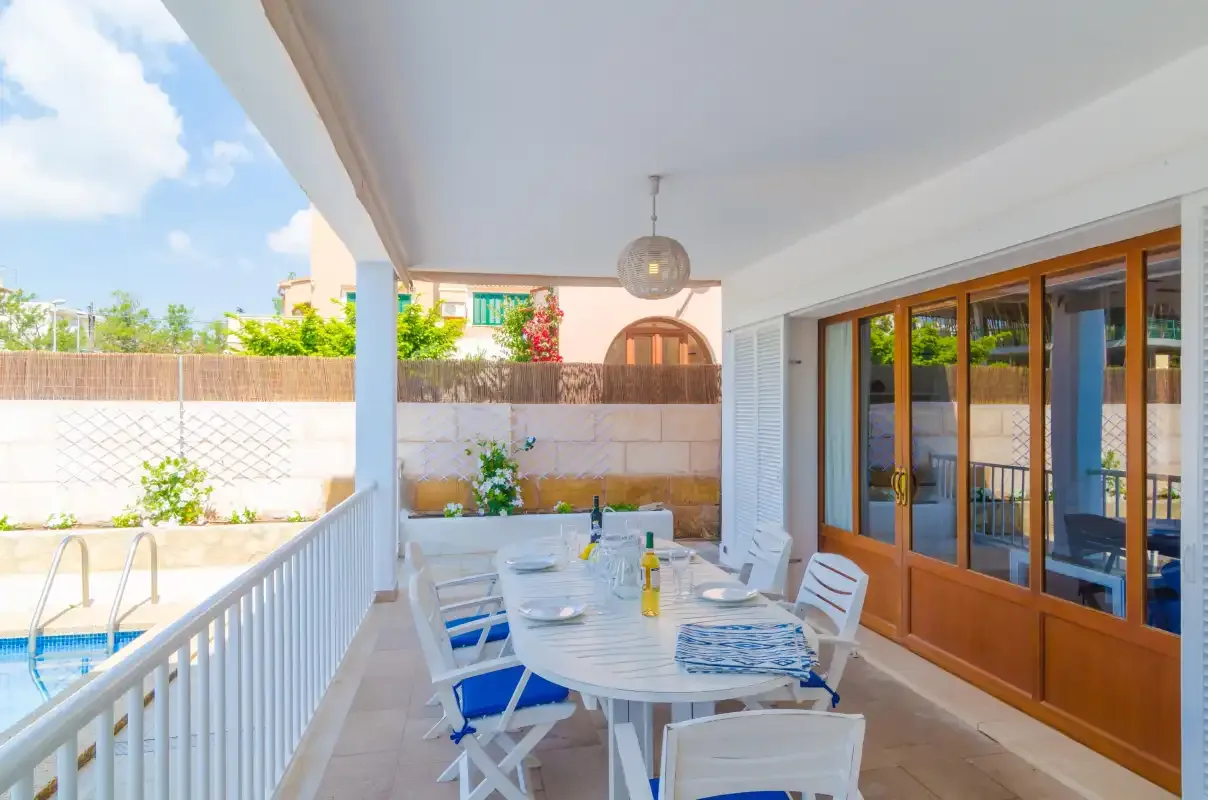 A covered patio has a dining table beside sliding glass doors and a swimming pool.