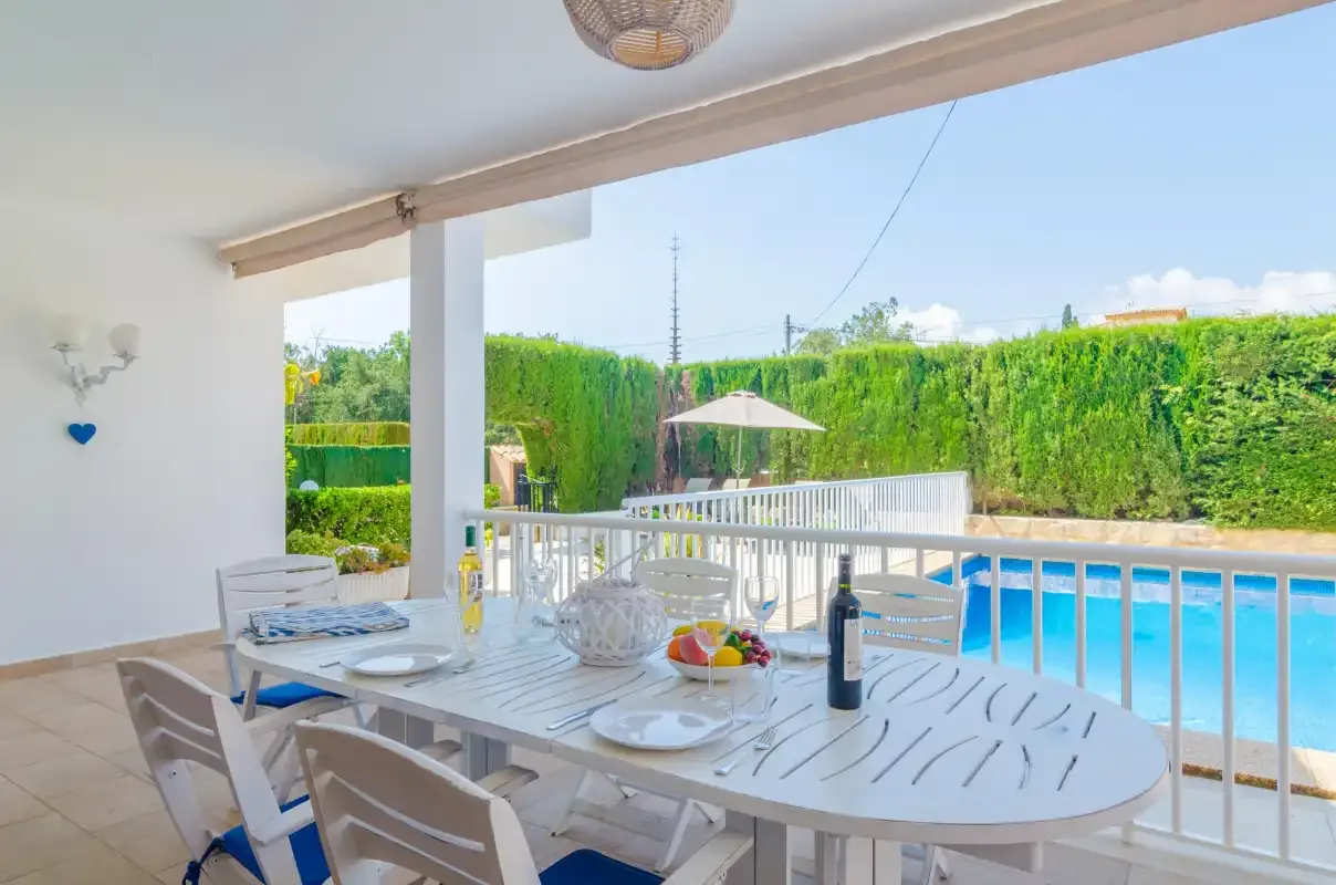 A covered outdoor dining area overlooks a swimming pool and green hedges.
