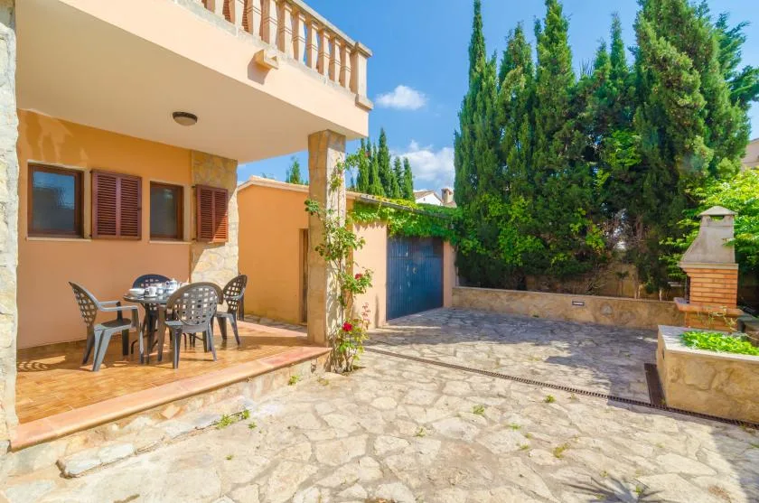 A sunny patio beside a house opens onto a paved courtyard with tall trees.