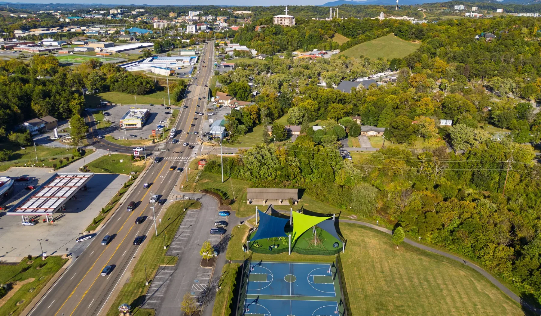 An aerial view of outdoor basketball courts beside a multi-lane road and wooded hills.