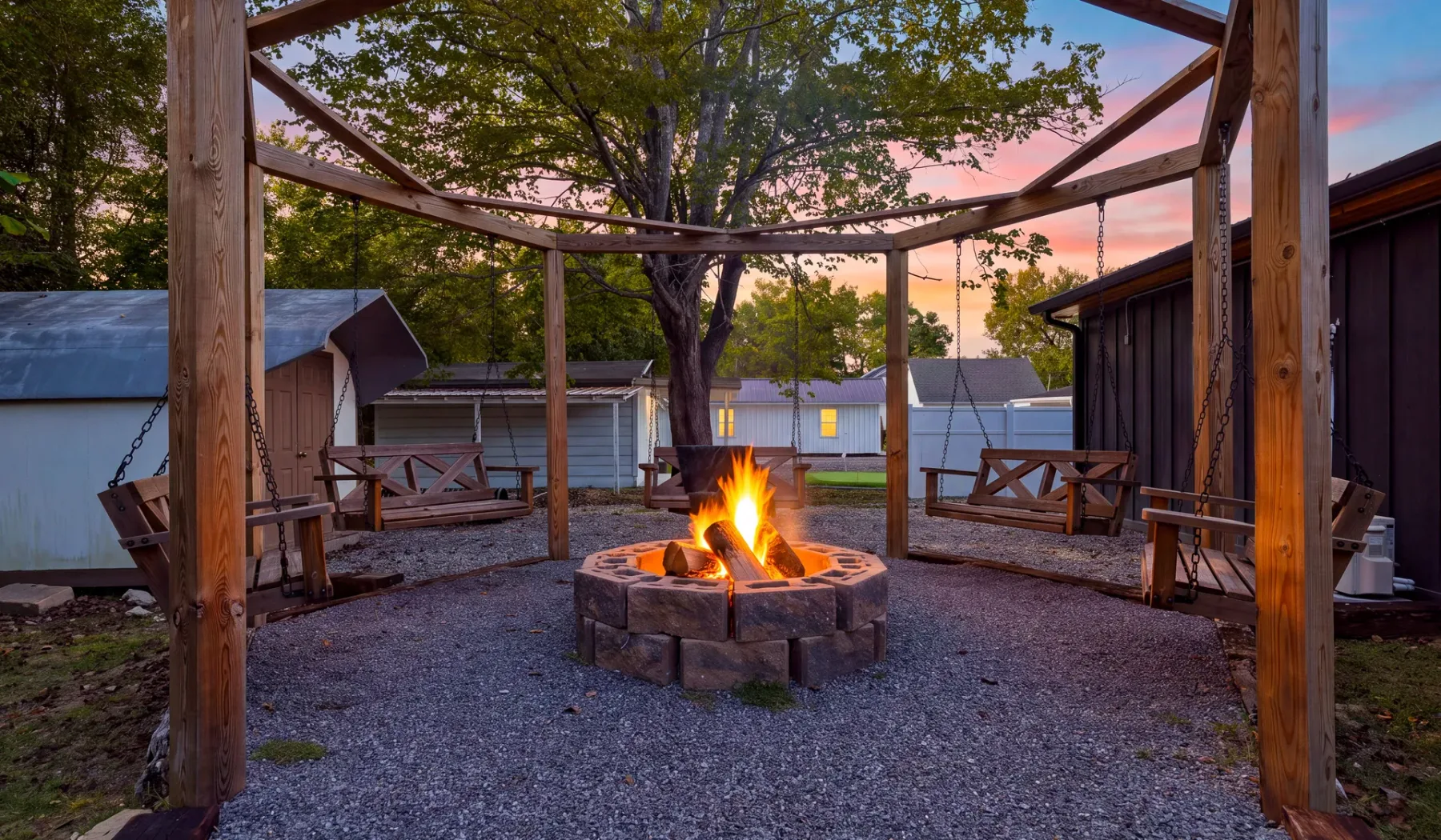 Un patio de grava con un fogón circular encendido bajo una pérgola de madera al atardecer.