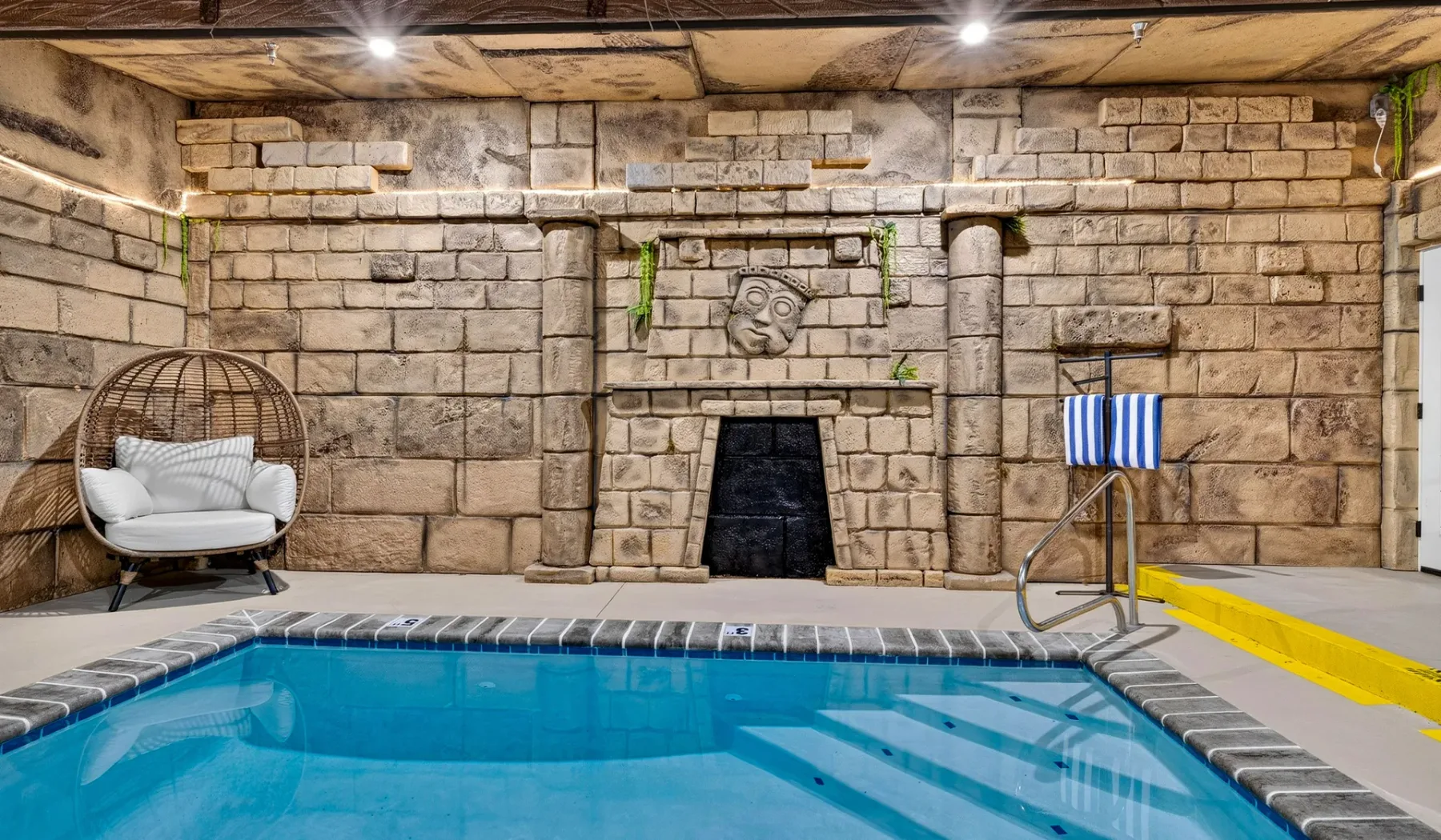 An indoor swimming pool sits beside a stone wall with a hanging chair.