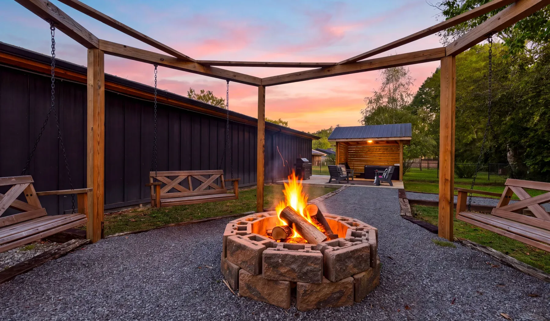 An outdoor fire pit glows beneath a wooden pergola at sunset.