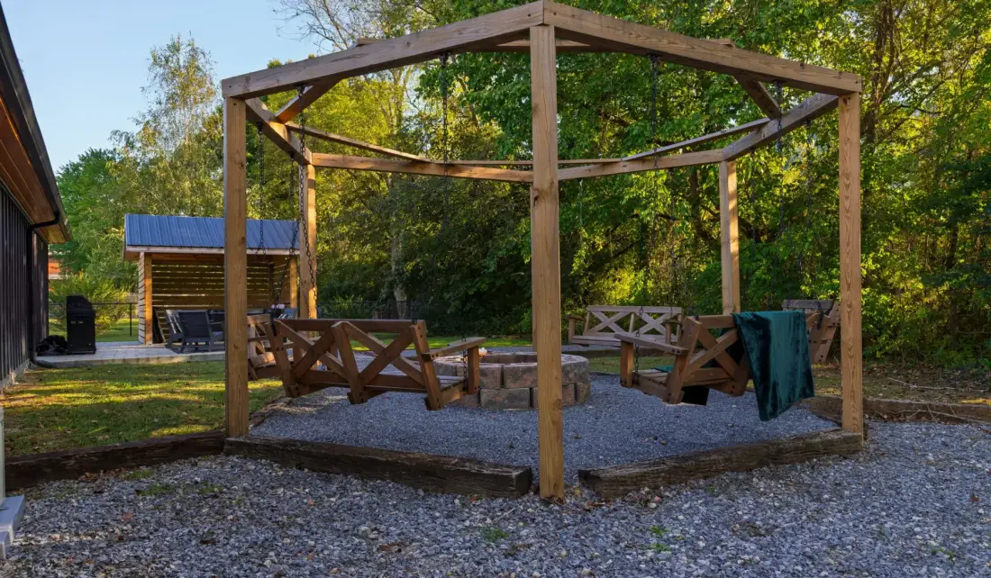 A wooden gazebo with benches stands in a gravel outdoor area.