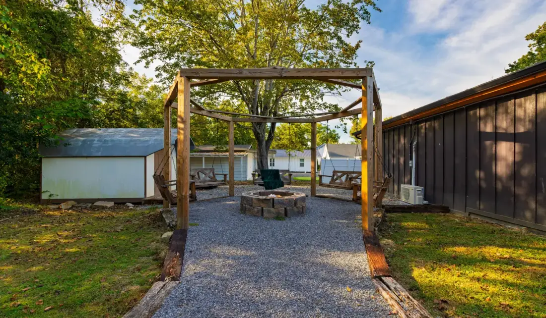 An outdoor gravel seating area sits beneath a wooden pergola, surrounded by trees.