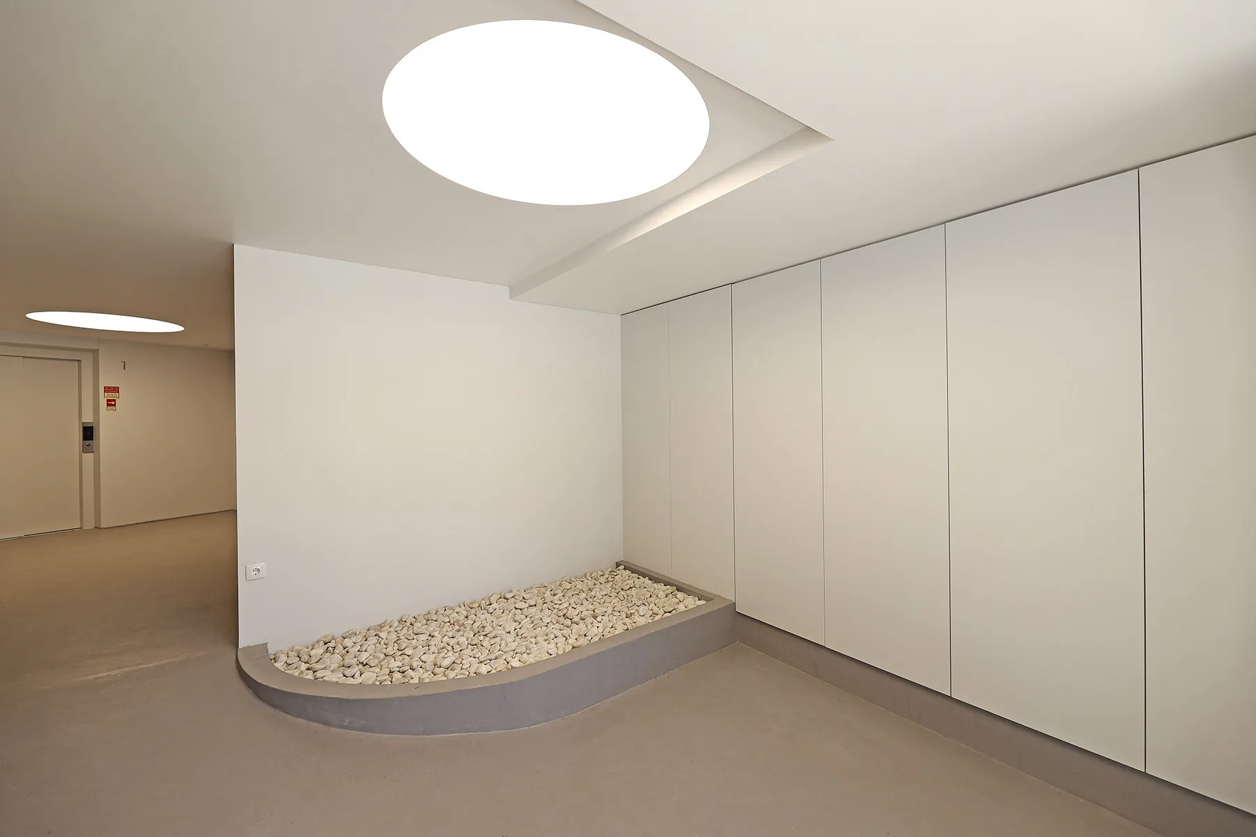 A minimalist indoor hallway with a curved, pebble-filled planter beside a row of wall panels.