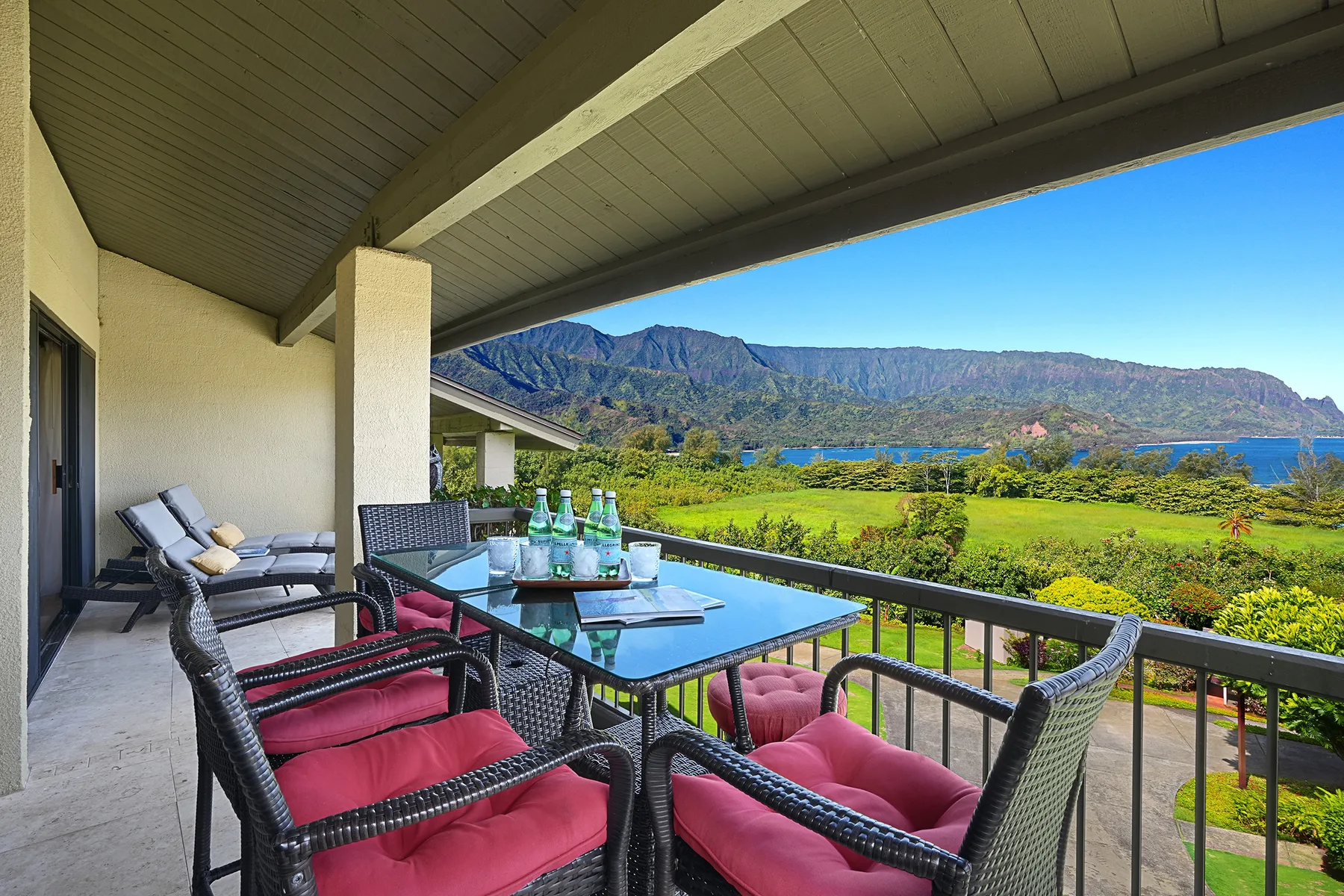 A covered patio has cushioned seating and a table overlooking greenery, water, and mountains.