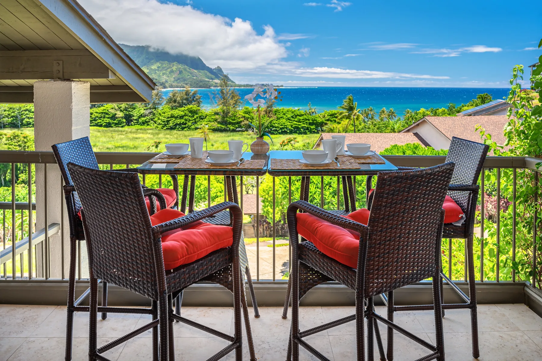 A balcony dining area with cushioned seating overlooks the ocean and green hills.