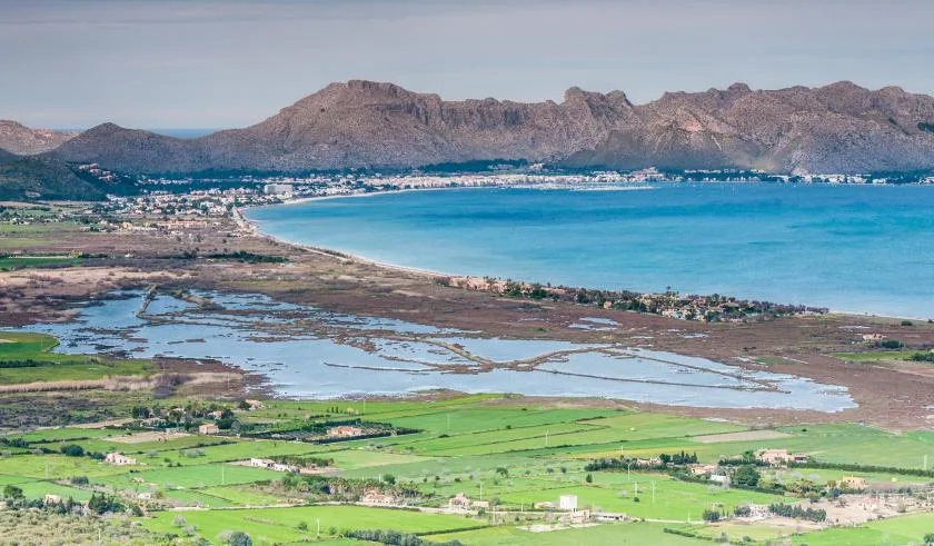 A coastal landscape with a turquoise bay, wetlands, and green fields beneath rocky mountains.