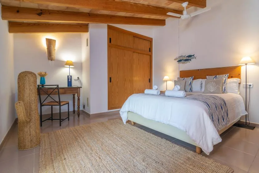 A bedroom with a made bed, wooden ceiling beams, and a desk in the corner.