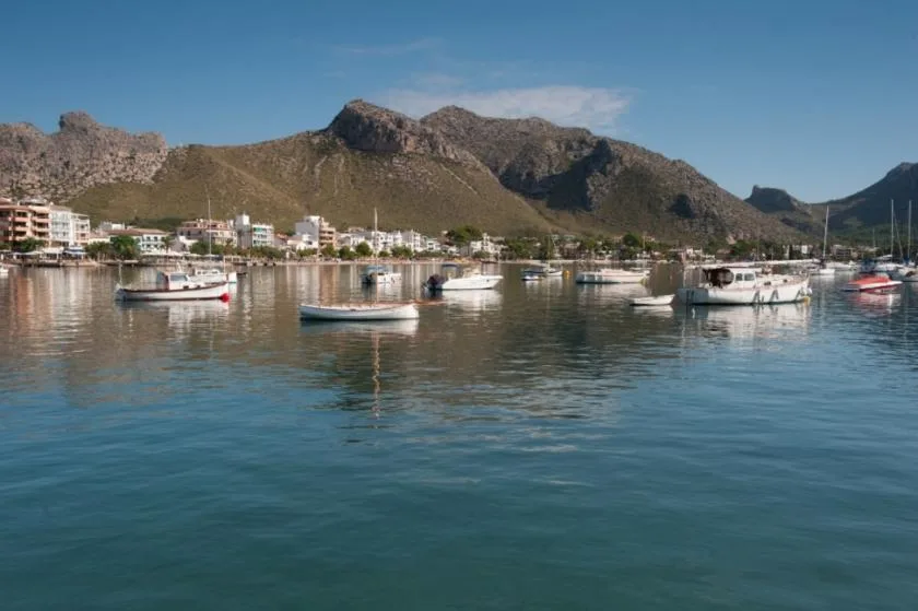 Boats float on calm harbor water, with a coastal town and mountains in the background.