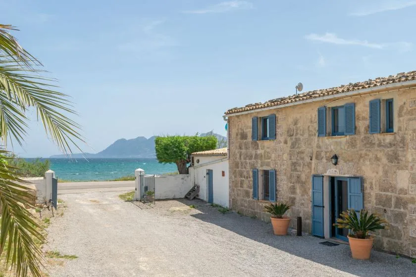 A stone building with blue shutters stands beside a gravel drive overlooking the sea.