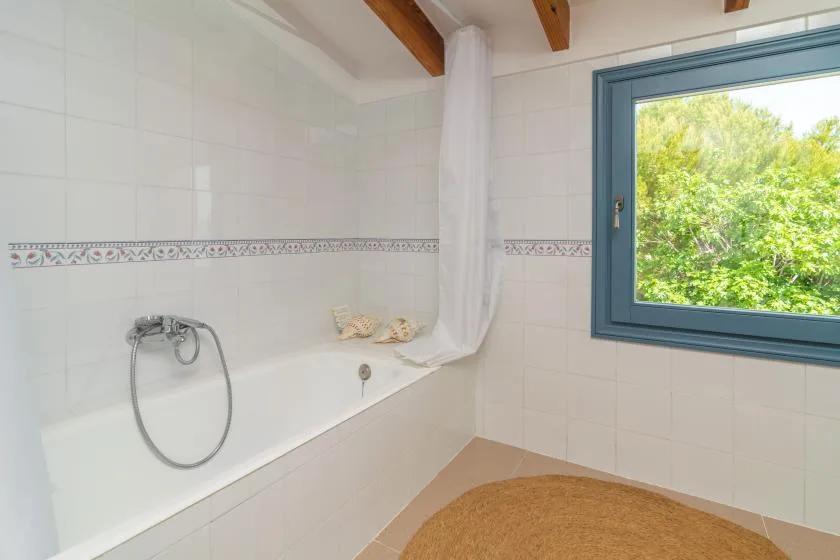 A tiled bathroom with a bathtub, shower hose, and window overlooking greenery.