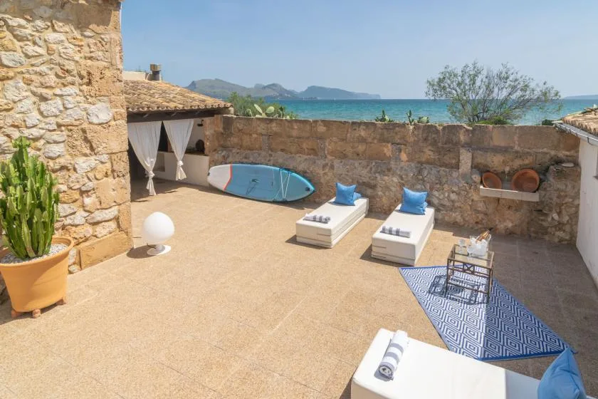 A sunny outdoor terrace with lounge seating, a surfboard, and a view of the sea.