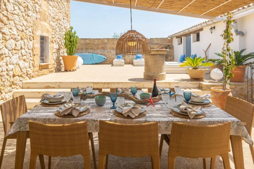 An outdoor dining table is set beneath a wooden pergola beside a sunlit pool.