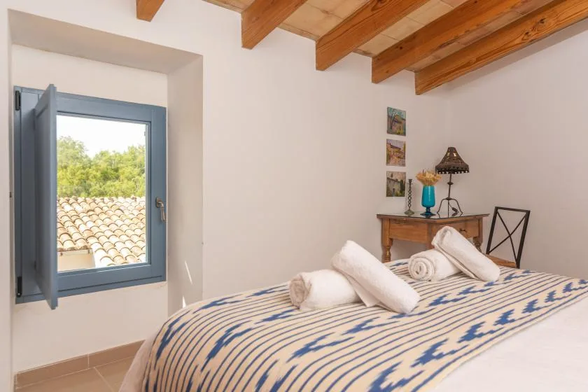 A bedroom with a patterned bed, an open window, and exposed wooden ceiling beams.