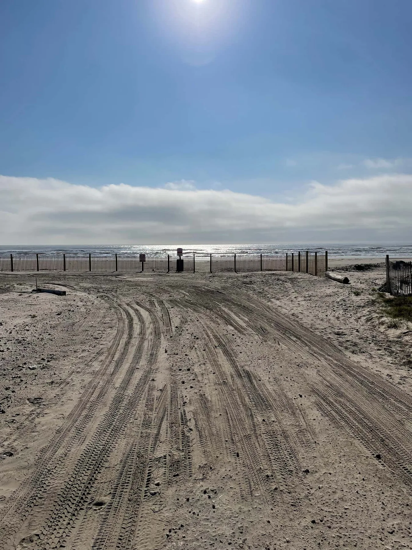 A sandy beach access track leads toward a fenced shoreline beneath a bright sky.