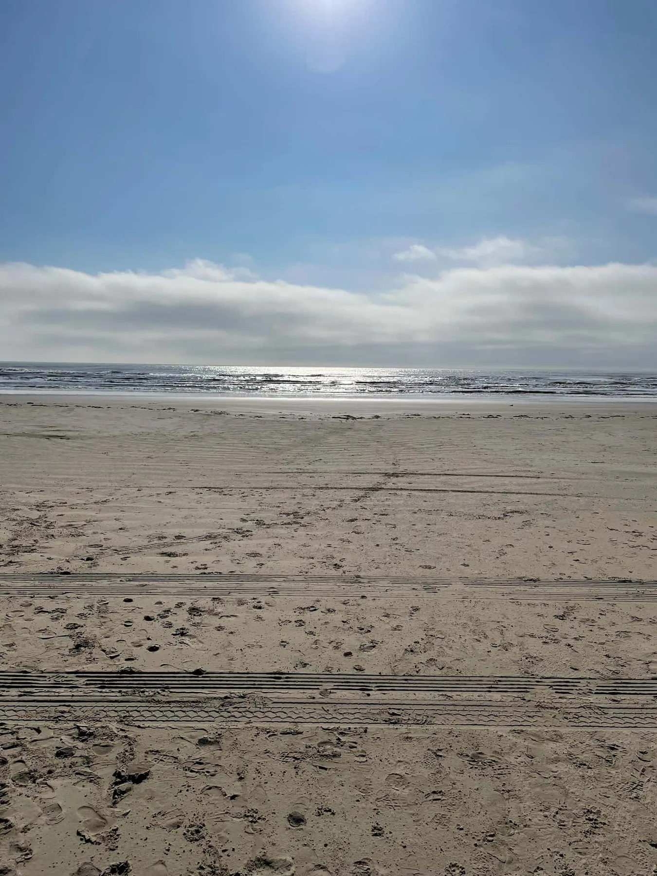A sandy beach stretches toward the ocean beneath a bright, clouded sky.