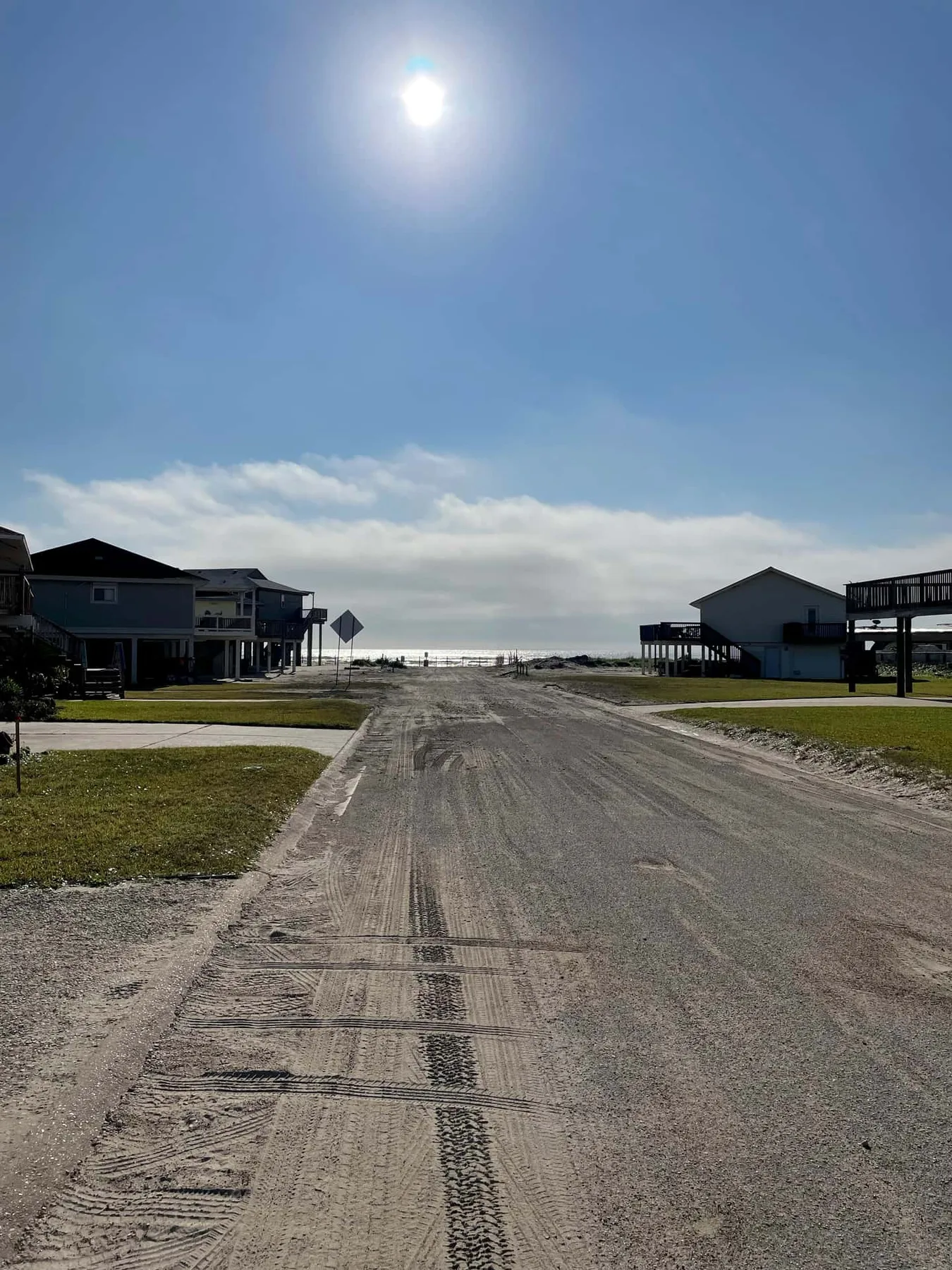 A sandy road runs between houses toward the water beneath a bright sky.
