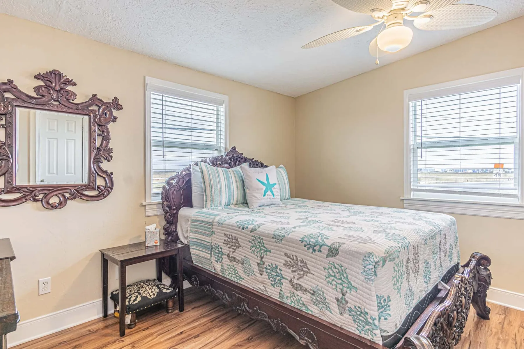 A bedroom with a bed, patterned bedding, and two windows with blinds.