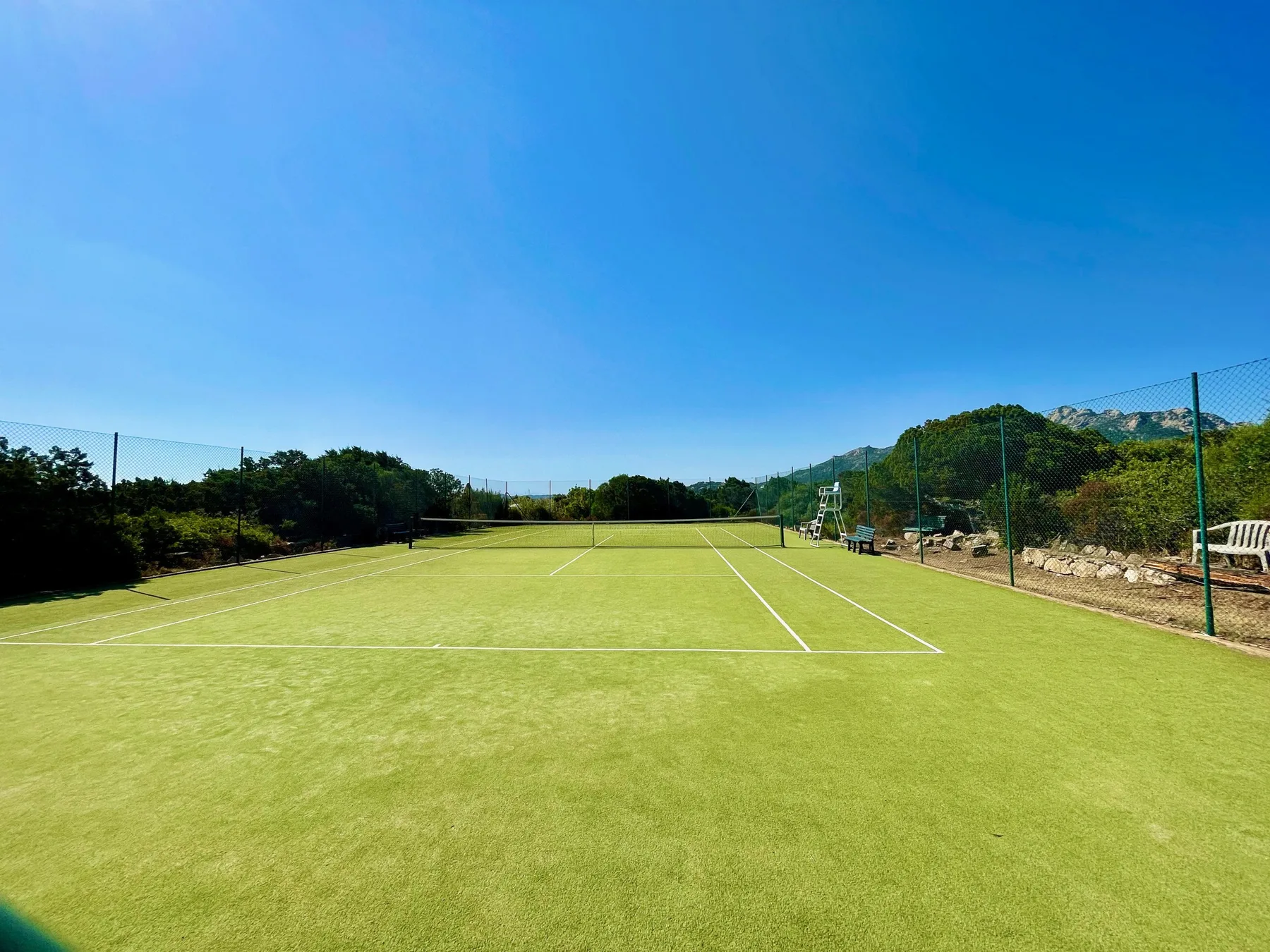 An outdoor tennis court with a grassy surface and surrounding fencing.