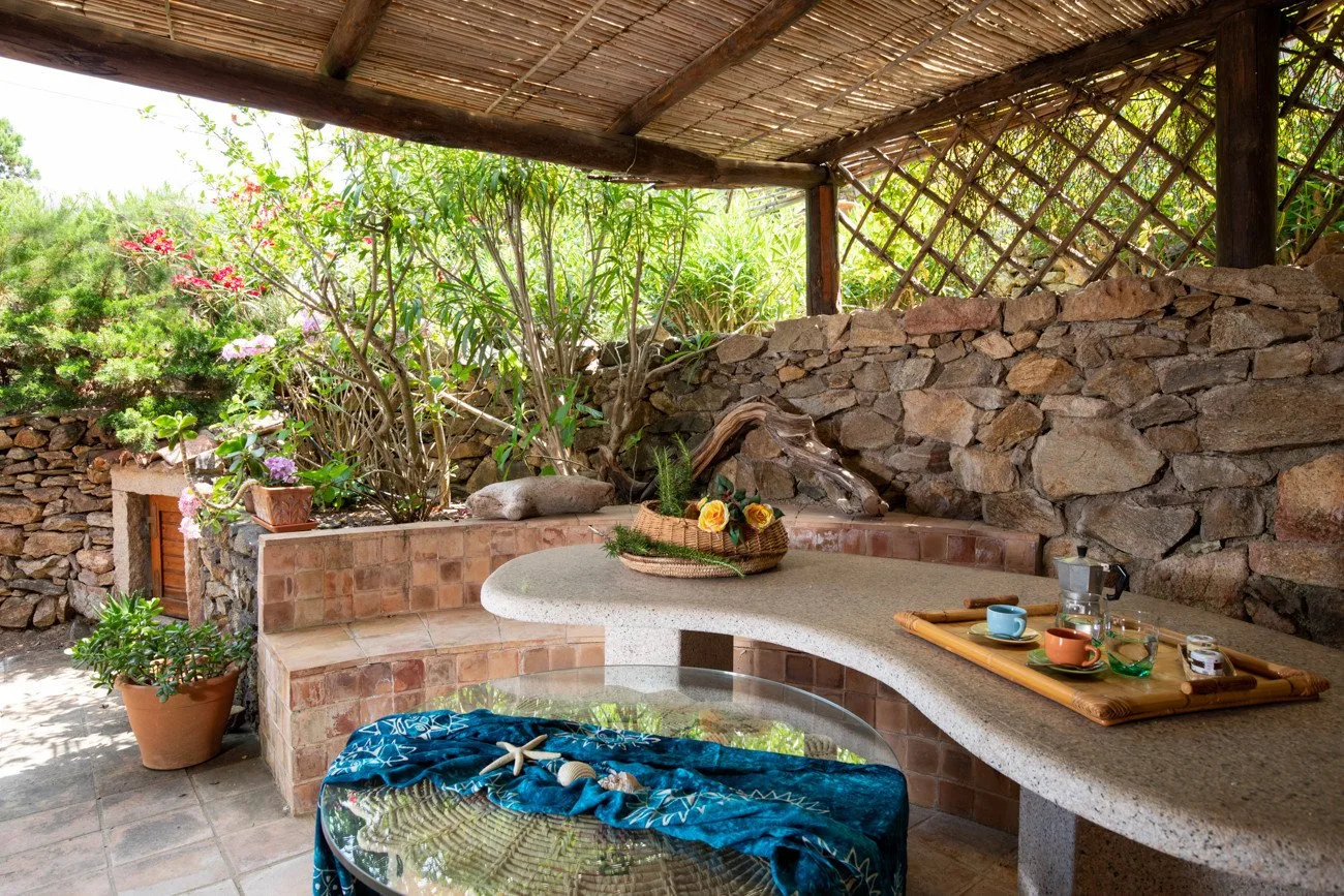 A covered outdoor seating area surrounds a tiled basin, with a stone wall and garden plants.