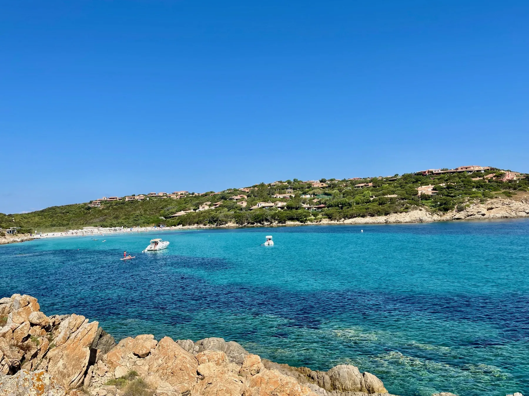 A turquoise bay stretches toward a green, rocky coastline under a clear blue sky.