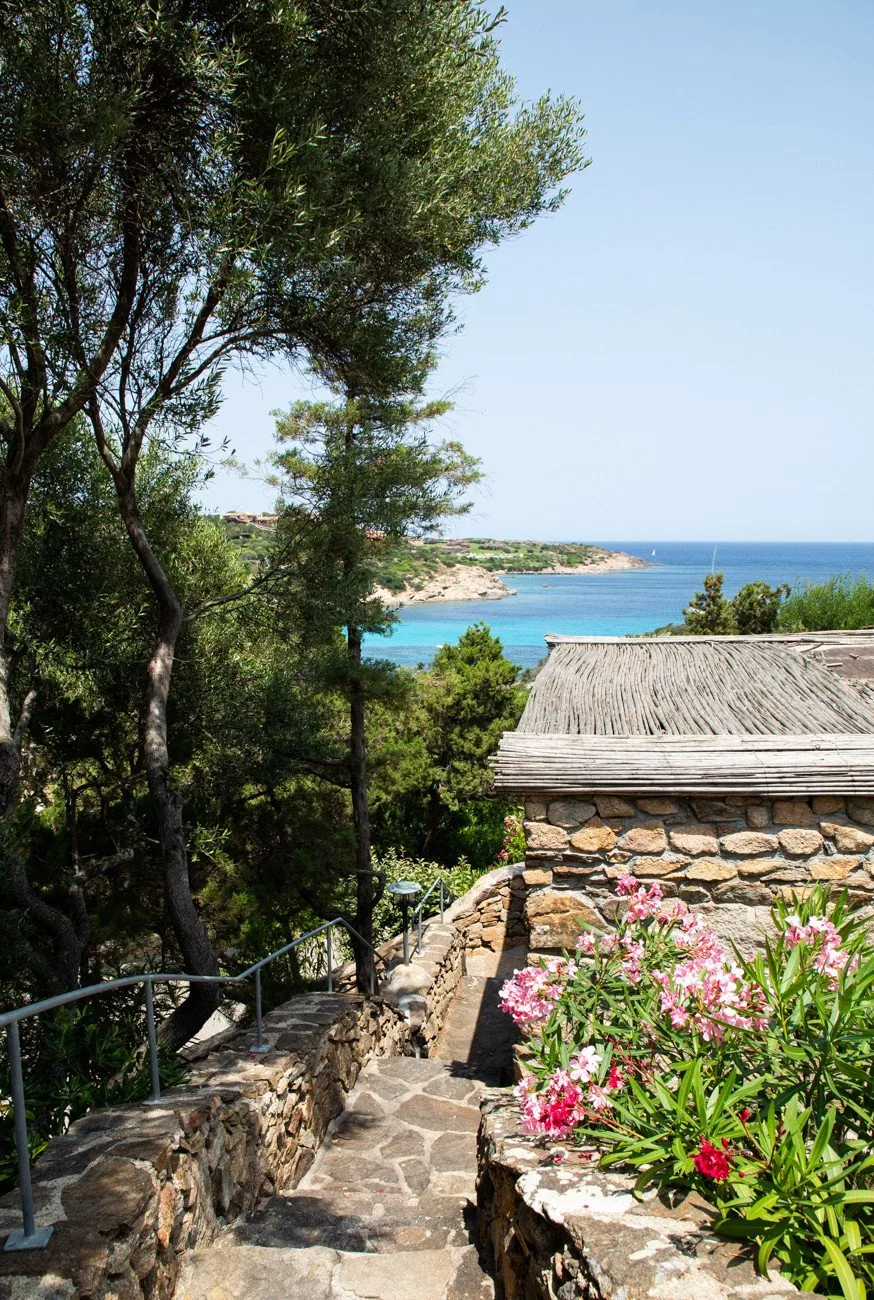 A stone path descends past a stone building toward a coastal bay.