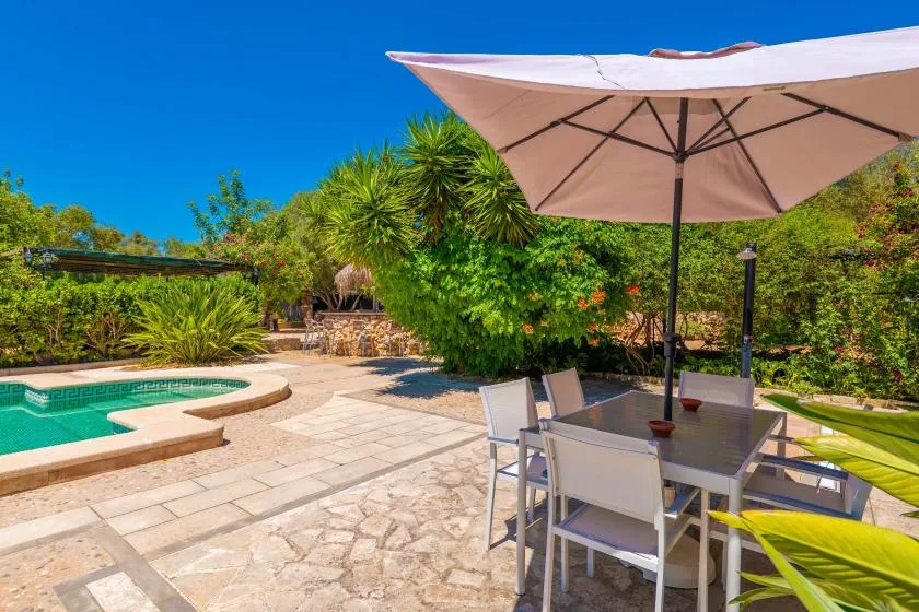 Un patio soleado tiene una mesa de comedor al aire libre junto a una piscina y un jardín.