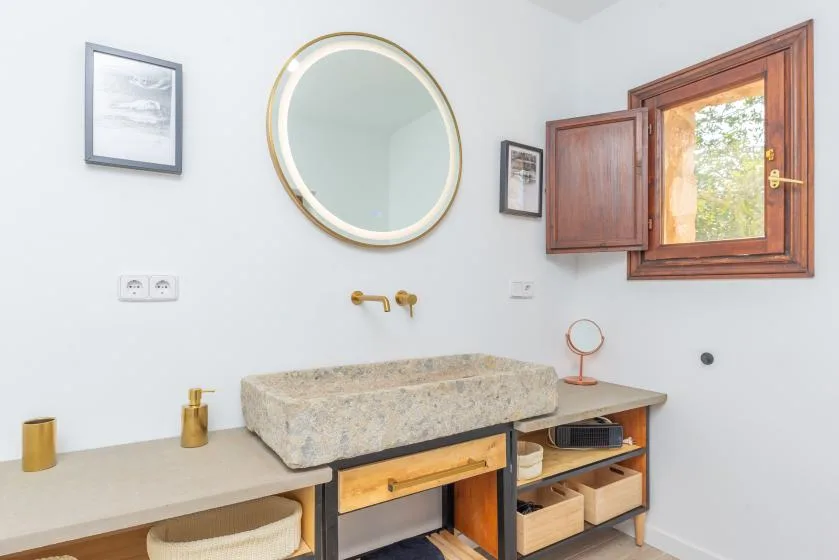 Un lavabo de baño luminoso con una pila de piedra, espejo redondo y ventana de madera.