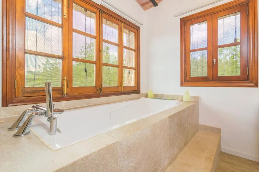 Una bañera empotrada está bajo ventanas con marcos de madera en un baño luminoso.