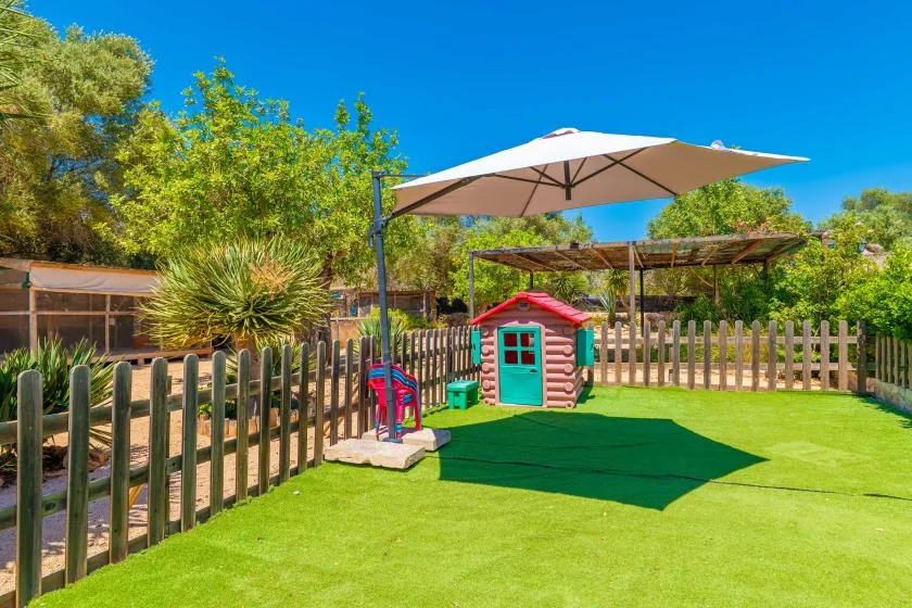 Una zona de juegos exterior cercada, con una casita bajo una sombrilla grande.