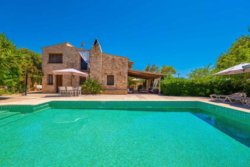 Una villa de piedra está junto a una piscina, con tumbonas y vegetación alrededor del patio.