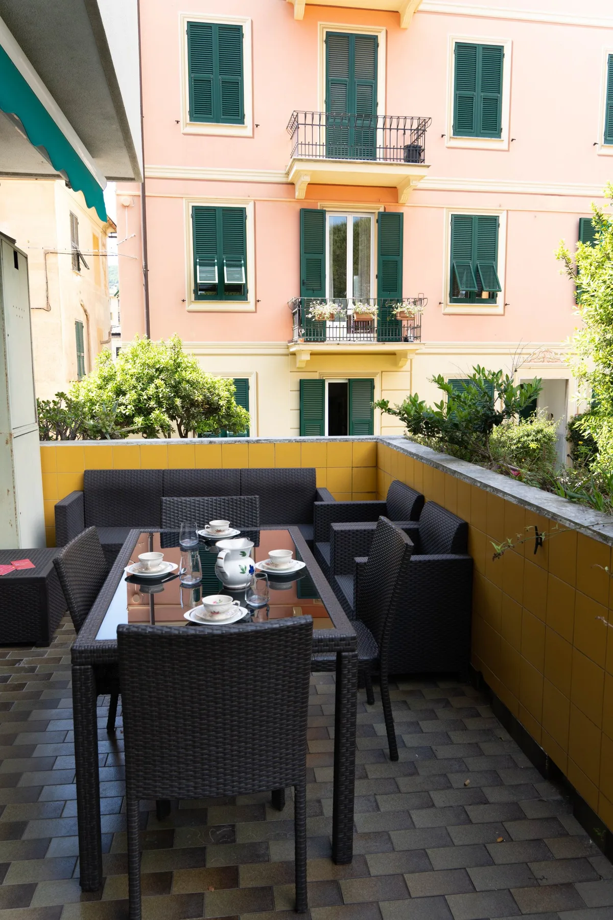 Una zona de comedor en una terraza exterior tiene asientos oscuros alrededor de una mesa junto a un paso embaldosado.