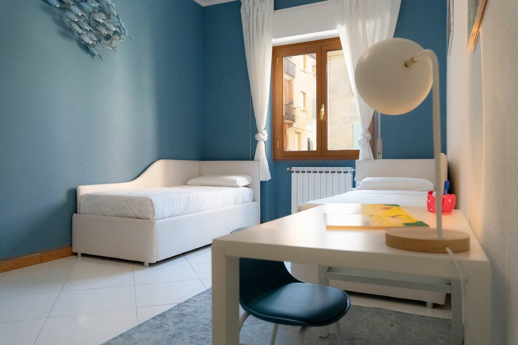 Un dormitorio de paredes azules con dos camas y un escritorio junto a la ventana.