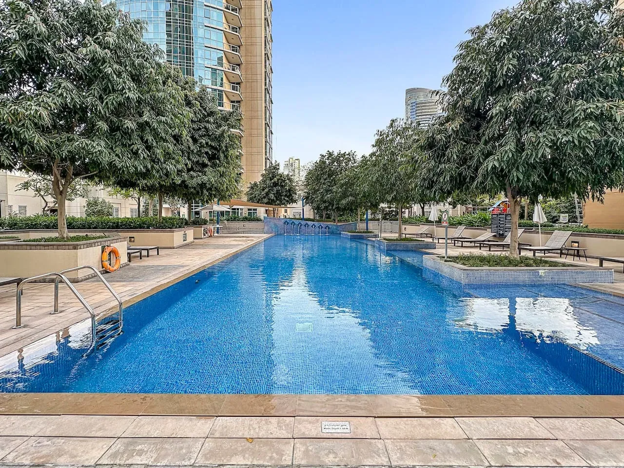 An outdoor swimming pool is surrounded by trees, paved deck, and tall buildings.